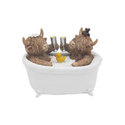 Highland Cow couple figurine sitting in a white roll-top bath clinking champagne flutes with a rubber duck, one cow wearing a hair clip