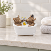 Two Highland cow figurines raising drinking flutes in a bubble-filled bathtub with a rubber duck, displayed on a bathroom shelf beside folded towels with tiles and a plant softly blurred behind.⠋
