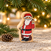 Wooden Father Christmas toy beneath Christmas tree branches beside a pinecone and warm fairy lights