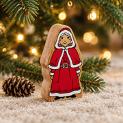 Wooden Mrs Claus toy beneath Christmas tree branches beside a pinecone and warm fairy lights