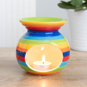 Rainbow Stripe Oil Burner, lifestyle shot of lit tealight glowing through oval cut-out in ceramic oil burner with bold rainbow stripes, wooden surface and plant in background