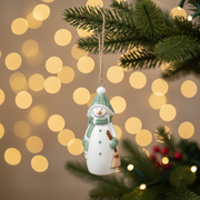 Hanging snowman Christmas tree decoration with sage green hat and mittens, suspended on a Christmas tree branch with warm golden fairy lights in the background