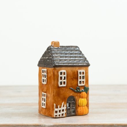 Pumpkin House ceramic tealight holder, amber two-storey ceramic house with dark grey slate-effect roof, amber chimney, small white-framed windows, grey door with white picket fence and stack of three pumpkins beside the entrance, white background