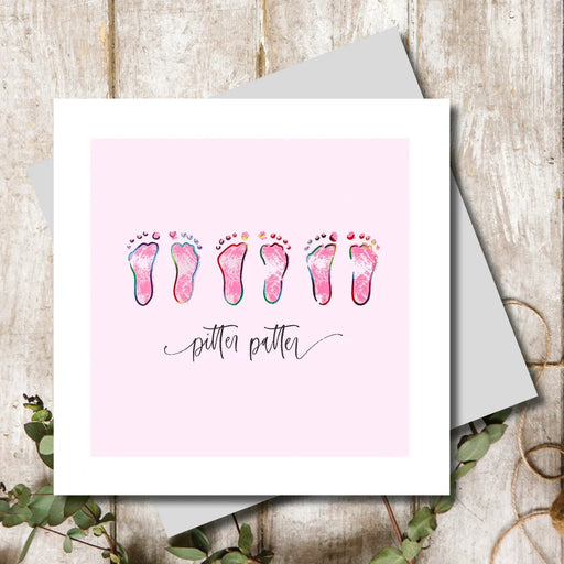 New baby girl card with three pairs of tiny rainbow foil baby footprints on a soft pink background with pitter patter sentiment in handwritten script, 155 x 155mm