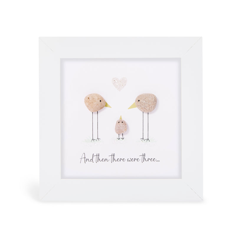 Pebble art framed plaque with two natural pebble birds facing each other with a tiny pebble chick between them and a small heart pebble above, with the sentiment "And then there were three...", white frame