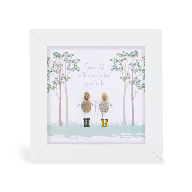 Pebble art framed plaque with two natural pebble figures standing hand in hand in a misty woodland scene, one in dark green wellies and one in yellow wellies holding a small flower, with the sentiment "Grow old with me, the best is yet to be", white frame