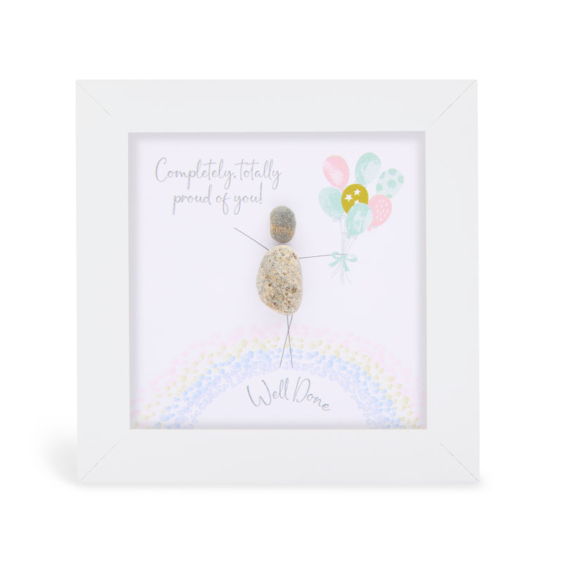Pebble art framed plaque with a natural pebble figure standing on a rainbow holding pastel balloons and the sentiment "Completely, totally proud of you! Well Done", white frame