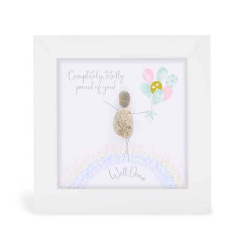 Pebble art framed plaque with a natural pebble figure standing on a rainbow holding pastel balloons and the sentiment "Completely, totally proud of you! Well Done", white frame