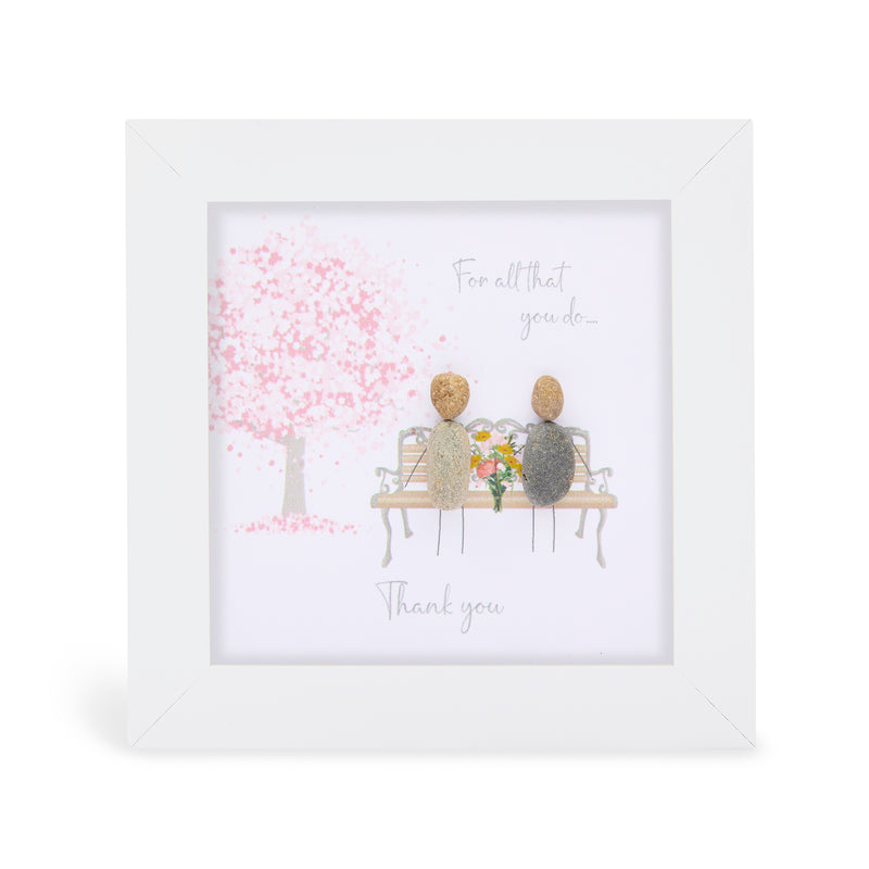 Pebble art framed plaque with two natural pebble figures sitting on a bench holding flowers beneath a blossom tree and the sentiment "For all that you do... Thank you", white frame