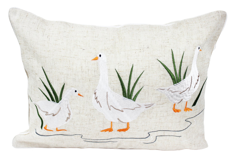 Puddles Embroidered Duck Cushion with three embroidered white ducks with orange beaks and feet in a puddle scene with green grass tufts on natural linen-mix background with white piped edge, 33 x 45 cm, on white background