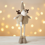 A tall fawn standing moose decoration with dark brown fabric antlers, a knitted beige pompom hat, googly eyes, a brown nose and a knitted scarf, standing against a warm golden Christmas background with soft bokeh fairy lights