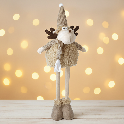 A tall fawn standing moose decoration with dark brown fabric antlers, a knitted beige pompom hat, googly eyes, a brown nose and a knitted scarf, standing against a warm golden Christmas background with soft bokeh fairy lights