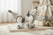 Sitting skating snowman decorations with grey striped bobble hats displayed on a wooden board with wrapped Christmas gifts and winter foliage