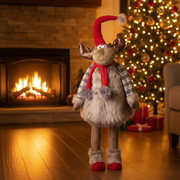 A large standing reindeer decoration with a red pompom hat, red scarf with fluffy pompoms and grey check arms, naturally placed in a warm cosy Christmas living room scene with soft golden ambient light

