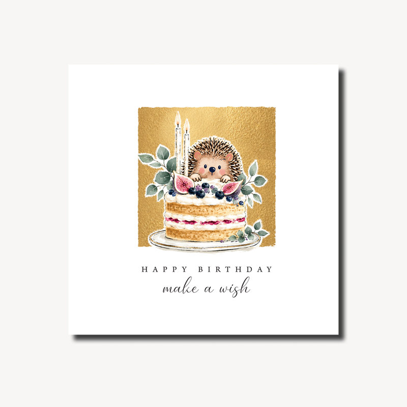 Happy birthday card featuring a hedgehog peeking from behind a decorated layered cake with berries, figs and candles, botanical leaves and gold foil background, 155 x 155mm