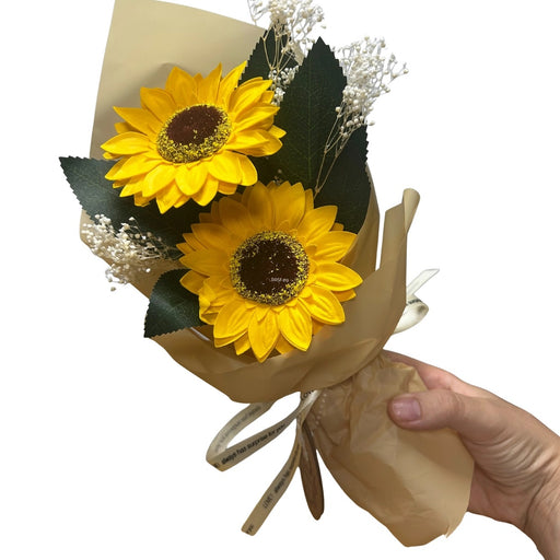 Sunflower Soap Flower Bouquet, hand holding a small bouquet of two soap petal sunflowers with gypsophila and dark green leaves wrapped in kraft paper with ribbon tie, white background