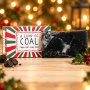 Neon Magpie Christmas Lump of Coal Vegan Soap Bar in red and green festive packaging styled on a pale wooden surface with coal and holly, warm golden bokeh background