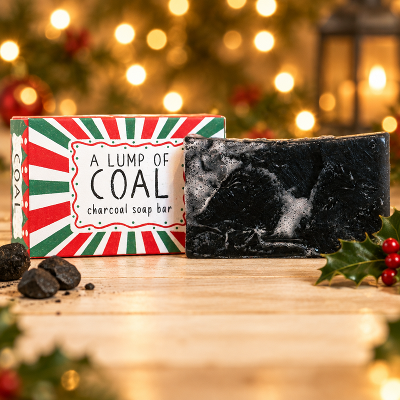 Neon Magpie Christmas Lump of Coal Vegan Soap Bar in red and green festive packaging styled on a pale wooden surface with coal and holly, warm golden bokeh background