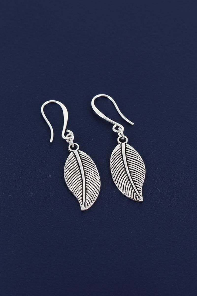 Silver plated leaf charm drop earrings with fine vein texture on hook fittings on a deep navy blue background