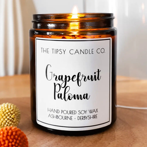 Tipsy Grapefruit Paloma Scented Candle lit in amber glass jar with warm golden glow on wooden surface