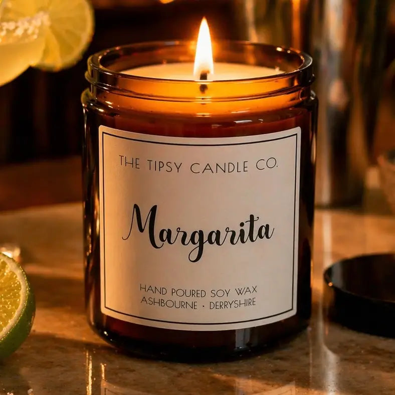 Tipsy Margarita Scented Candle lit in amber glass jar with lime and lemon on marble surface