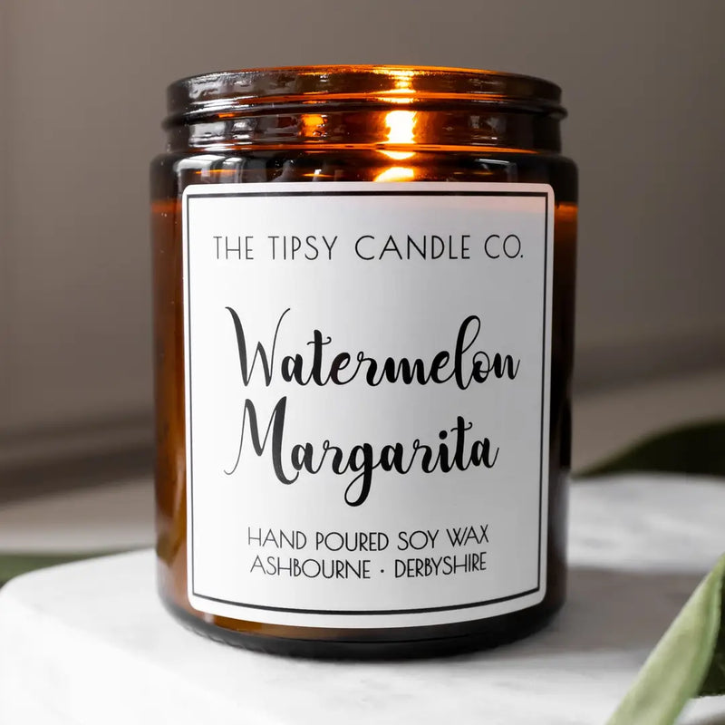 Tipsy Watermelon Margarita Scented Candle in amber glass jar on white marble surface with green foliage