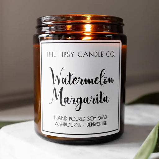 Tipsy Watermelon Margarita Scented Candle in amber glass jar on white marble surface with green foliage