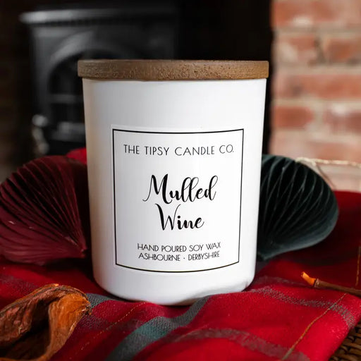Tipsy Mulled Wine Scented Candle in white matte glass jar with wooden lid styled by a fireplace on tartan fabric