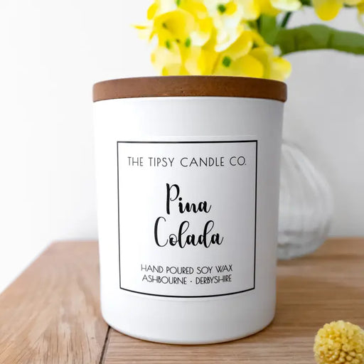 Tipsy Piña Colada Scented Candle in white matte glass jar with wooden lid on wooden surface with yellow flowers