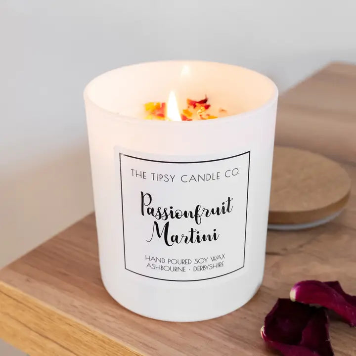 Tipsy Passionfruit Martini Scented Candle lit in white glass jar with dried botanicals on wax surface on wooden surface with rose petals