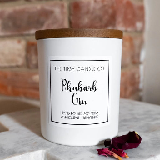 Tipsy Rhubarb Gin Scented Candle in white matte glass jar with wooden lid on marble surface against brick wall with rose petals