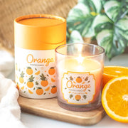 Orange Scented Candle, lifestyle shot of lit candle in clear glass jar alongside illustrated cylindrical gift box on wooden board with fresh orange slices, plant in background