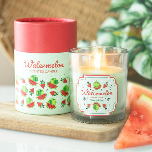 Watermelon Scented Candle, lifestyle shot of lit candle in clear glass jar with watermelon label alongside illustrated cylindrical gift box, wooden board and fresh watermelon slice in background