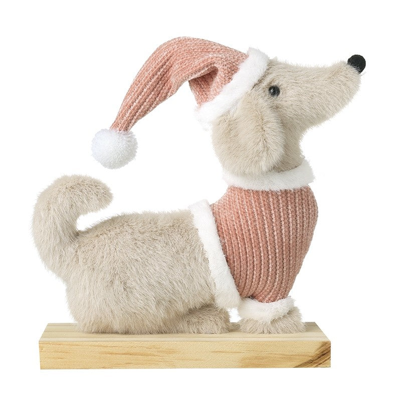 A soft beige sausage dog ornament wearing a dusky pink ribbed jumper with white trim and a matching slouchy Christmas hat with a white pompom, sitting on a wood-effect base, on a white background