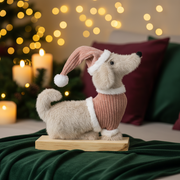 A soft beige sausage dog ornament wearing a dusky pink ribbed jumper with white trim and a matching slouchy Christmas hat with a white pompom, sitting on a wood-effect base, styled in a warm cosy Christmas setting with golden bokeh fairy lights and subtle evergreen hints in the background