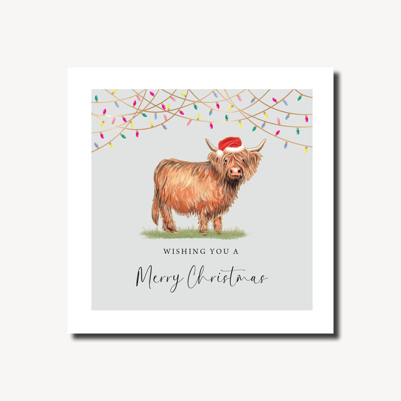 Merry Christmas card featuring a fluffy Highland cow in a Santa hat standing beneath strings of colourful fairy lights on a soft grey background, foil finish, 155 x 155mm