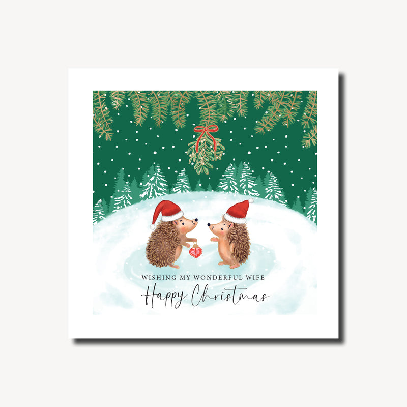 Christmas card for a wonderful wife featuring two hedgehogs in Santa hats facing each other under a sprig of mistletoe in a snowy winter scene with pine branches and fir trees, gold foil finish, 155 x 155mm