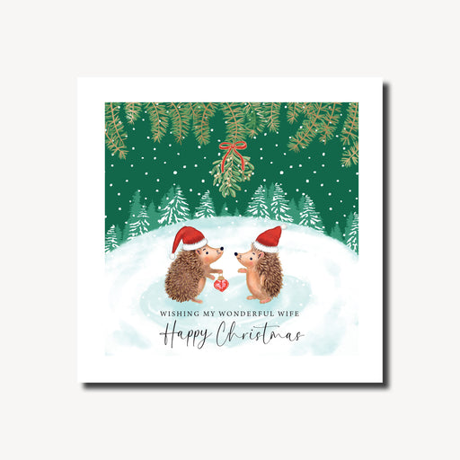 Christmas card for a wonderful wife featuring two hedgehogs in Santa hats facing each other under a sprig of mistletoe in a snowy winter scene with pine branches and fir trees, gold foil finish, 155 x 155mm