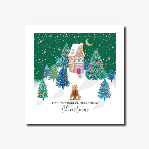 Christmas card for a wonderful husband featuring two hares sitting together in the snow gazing at a warmly lit pink house with snow-topped roof, surrounded by decorated fir trees under a deep green starry night sky, gold foil finish, 155 x 155mm