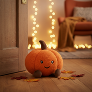 A plush orange pumpkin doorstop with a fluffy sherpa texture, smiley face, dark brown stem and small dark brown feet, displayed in a warm cosy autumnal indoor setting with golden amber tones and soft bokeh fairy lights in the background