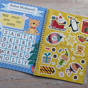 Big Sticker Activity Book Christmas Santa, open spread showing festive wordsearch on left and yellow sticker sheet on right with Santa's sleigh, penguin, elf, robin, teddy bear, gifts and baubles, wooden surface