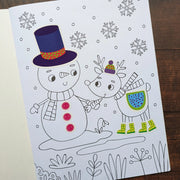Dress Me Up Snowman Colouring and Activity Book, lifestyle shot showing completed snowman colouring page with top hat and button stickers applied alongside reindeer in boots, wooden surface