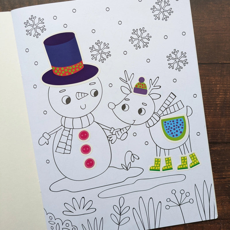 Dress Me Up Snowman Colouring and Activity Book, lifestyle shot showing completed snowman colouring page with top hat and button stickers applied alongside reindeer in boots, wooden surface