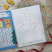 Sparkly Santa Gem Art Activity Book, lifestyle shot showing word search activity on left and children watching snow colouring page on right with gem sticker sheet, wooden surface