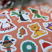 Sparkly Santa Gem Art Activity Book, lifestyle shot showing sheet of colourful Christmas character stickers including Mrs Claus, penguin, elf, robin, teddy bear and wreath on orange background