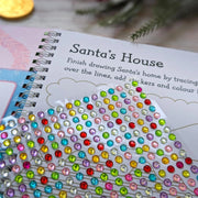Sparkly Santa Gem Art Activity Book, lifestyle shot showing sheet of multicoloured gem stickers alongside open Santa's House tracing and colouring page, festive props