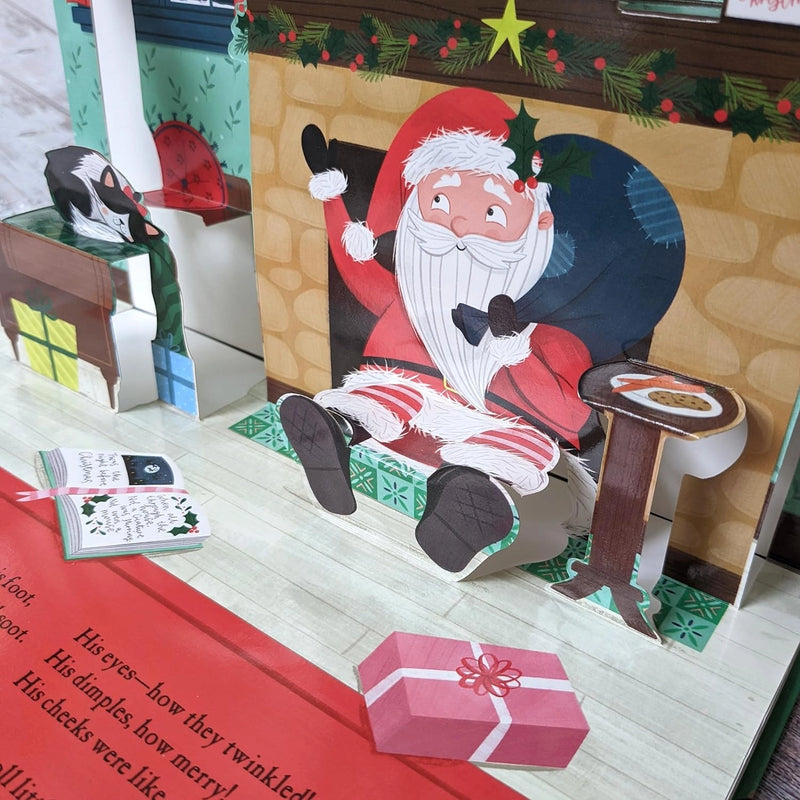 The Night Before Christmas Pop-Up Book open spread showing Santa seated by the fireplace with a plate of cookies on a side table and a sleeping cat to the left — 3D pop-up scene
