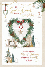 Special Couple Christmas card featuring a heart-shaped foliage wreath with pinecones, berries, a ribbon bow and hanging star, alongside little houses, Christmas trees and JOY lettering.