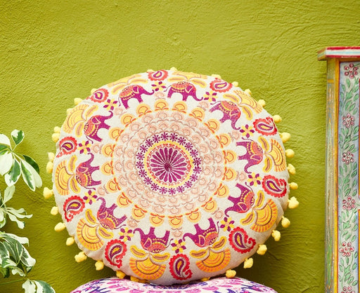 Round hand crafted elephant mandala cushion with pom-pom edging, showing intricate embroidered mandala design with a ring of elephants and floral motifs, styled against a green wall