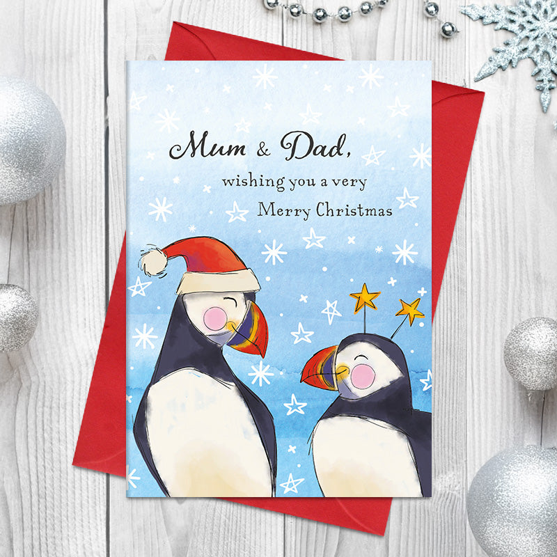 Mum and Dad Merry Christmas card featuring two illustrated puffins, one wearing a Santa hat and the other star deely boppers, surrounded by stars and snowflakes